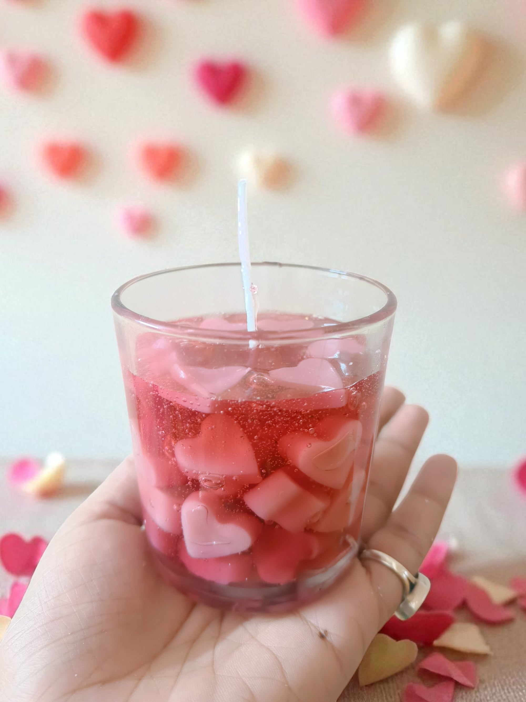 Hand holding a small glass filled with pink heart-shaped candle and a lit candle, with a blurred heart-patterned background.  Heart Latte candle Candelevate